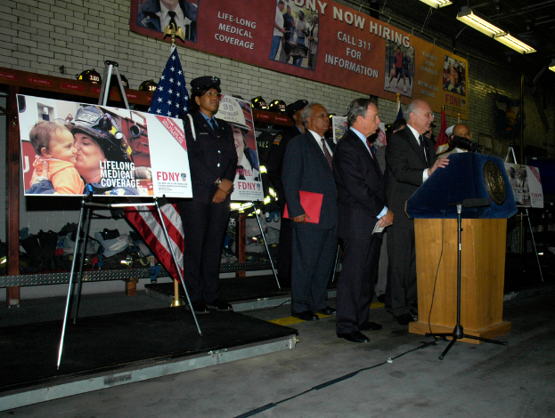 Eine Gruppe von Menschen steht neben einem Podium mit Mikrofonen, Schildern auf Ständern, Flaggen und Schildern an der Wand, alles unter hellen Scheinwerfern.