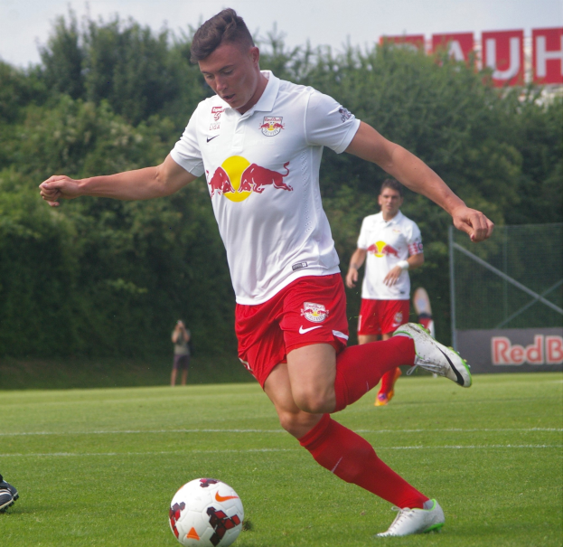 Ein Mann in einem Fussballtrikot kickt einen Ball auf einem grünen Feld mit Bäumen und einem klaren blauen Himmel im Hintergrund, mit einem Schild mit der Aufschrift "RB Leipzig" sichtbar.