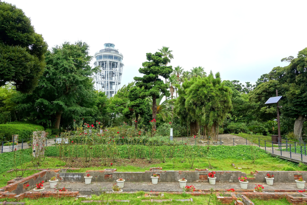 Ein Park mit einem Turm im Hintergrund, umgeben von grünem Gras, Pflanzen, Bäumen und verstreuten Blumentöpfen, mit einer Straße mit Geländern und einer Pfostenbefestigung unter einem sichtbaren Himmel.