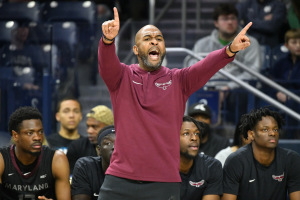 Ein Mann in einem roten Hemd und schwarzen Shorts steht am Spielfeldrand einer Basketballhalle und diskutiert mit einer Gruppe von Menschen, einige sitzen, andere stehen.