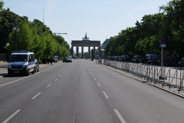 Ein Polizeifahrzeug parkt am Straßenrand vor dem Brandenburger Tor in Berlin, Deutschland, mit Barrieren, Hinweistafeln, Bäumen und Laternenmasten im Hintergrund und einer bewölkten Himmel.