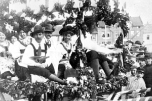 Eine Gruppe von Menschen in traditioneller bayrischer Tracht fährt auf einemblattgeschmückten Festwagen in einem Umzug mit, einige halten Musikinstrumente, mit Gebäuden und einem klaren Himmel im Hintergrund, dargestellt in Schwarz-Weiß.