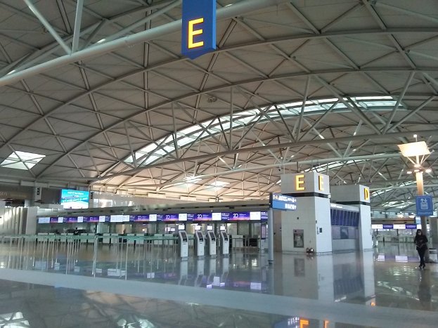 Leerer Flughafen-Terminal mit Menschen, die an Texttafeln und Maschinen sowie strukturellen Elementen wie Metallstangen und Lampen vorbeigehen.