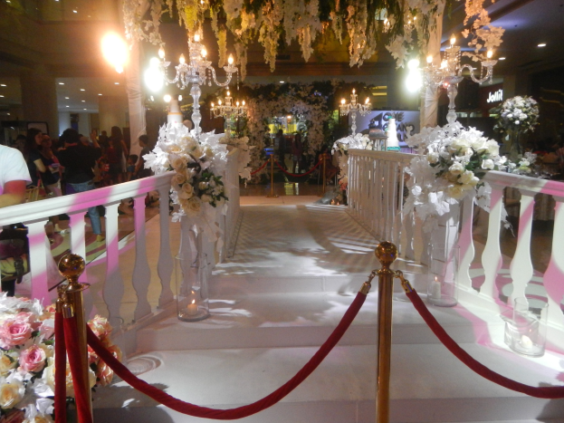 Eine Hochzeitszeremonie in einem Einkaufszentrum mit weißem Teppich, dekorativen Geländern, Blumenarrangements und Kronleuchtern, bei der viele Menschen anwesend sind, einige Taschen tragen, unter einer beleuchteten Decke.