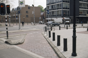 Eine Straßenszene in der Stadt mit einem geparkten Fahrrad, fahrenden Fahrzeugen, Fußgängern, Pfählen, Verkehrsampeln, Schildern, Bäumen, Gebäuden mit Fenstern und einem klaren blauen Himmel.
