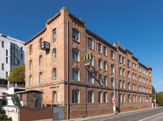Ein großes McDonald's-Restaurantgebäude aus Backstein an einer Straßenecke in Berlin, Deutschland, mit umgebender Straßeninfrastruktur, Fahrzeugen, Grünflächen und einem bewölkten Himmel.