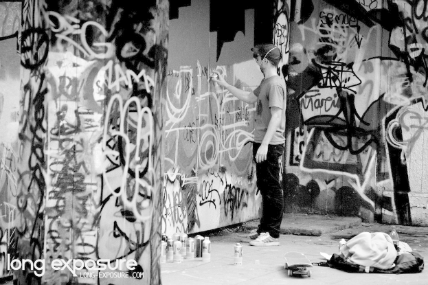 Ein Mann steht drinnen und h√§lt eine Flasche in der Hand, mit verstreuten Gegenst√§nden wie anderen Flaschen, einem Skateboard, einer Wasserflasche und Klamotten auf dem Boden, Graffiti an den W√§nden und Text in der linken unteren Ecke.