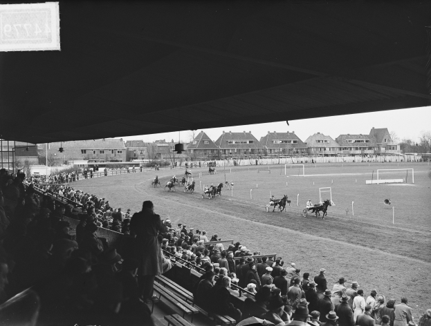Schwarz-weiß-Foto eines Pferderennens in einer Stadion mit Zuschauern auf Bänken und Jockeys, die Pferde auf der Bahn reiten, mit Gebäuden, Bäumen und Pfählen im Hintergrund unter einem klaren Himmel.