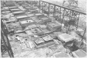 Ein Schwarz-Weiß-Foto einer Baustelle mit einer Brücke im Hintergrund, das Gebäude, Metallstangen und andere Gegenstände zeigt, mit Text unten.