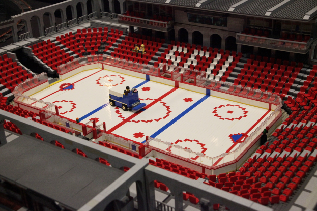 Ein Lego-Modell einer Eishockeyarena in einem Stadion mit einem Fahrzeug auf dem Eis, umgeben von sitzenden Figuren, Geländern und Treppen am Boden.