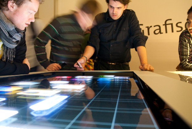 Ein Mann und ein Junge, beide in schwarzen Hemden, sehen auf einen Bildschirm; der Junge trägt einen Schal um den Hals, und ein cremefarbener Banner ist im Hintergrund zu sehen.