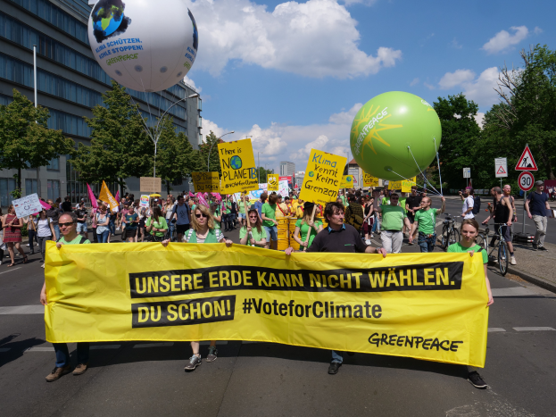 Eine Gruppe von Menschen marschiert mit einer gelben "Wähle für das Klima"-Schleife, Ballons, Schildern, Fahrrädern, Bäumen, Laternenpfählen, Gebäuden und einem klaren blauen Himmel die Straße entlang.