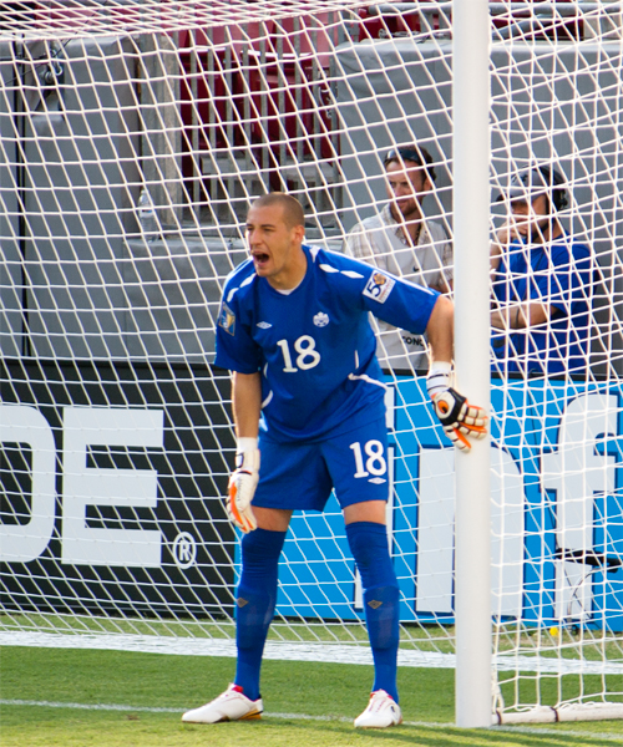 Ein Fußballspieler steht mit ausgestreckten Armen vor einem Tor, trägt einen Sportdress und Schuhe, mit drei Personen, einem Netz und einer Tafel mit Schrift im Hintergrund.