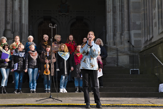 Eine Gruppe von Menschen, einige mit Musikinstrumenten, steht vor einem Gebäude mit Stufen und Geländern, während eine Frau in der Mitte in ein Mikrofon singt, umgeben von Mikrofonstativen.