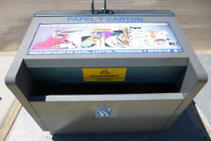 Ein Metallmüllcontainer am Straßenrand mit einem "Papel y Carton"-Plakat, im Hintergrund fahren Fahrzeuge.