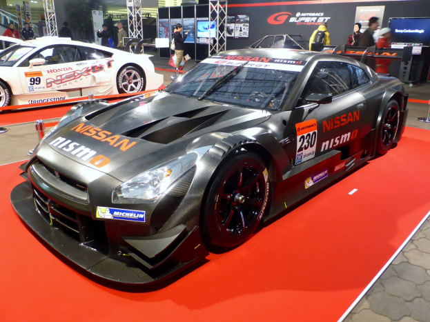 Ein Nissan GT-R Nismo GT3 Rennwagen auf einer Autauschstellung umgeben von Menschen, Absperrpoller mit Bändern, Schilder, Fernseher, Metallrahmen und Deckenleuchten und -lüfter.