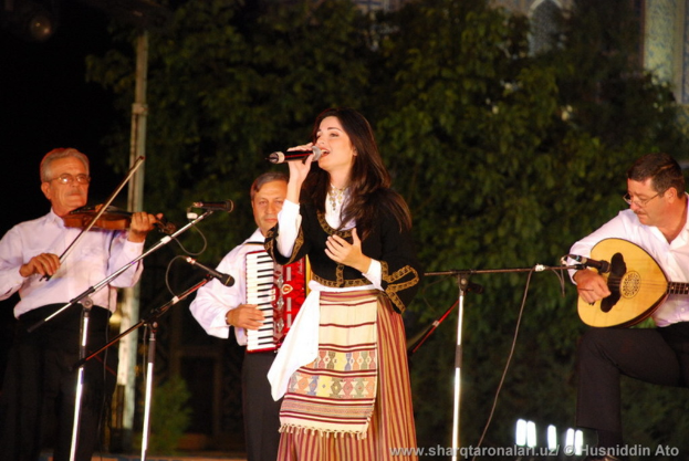 Eine Frau in einem Kleid singt in ein Mikrofon vorne, während drei Männer in der Nähe Musikinstrumente spielen und Bäume mit Lichtern im Hintergrund zu sehen sind.