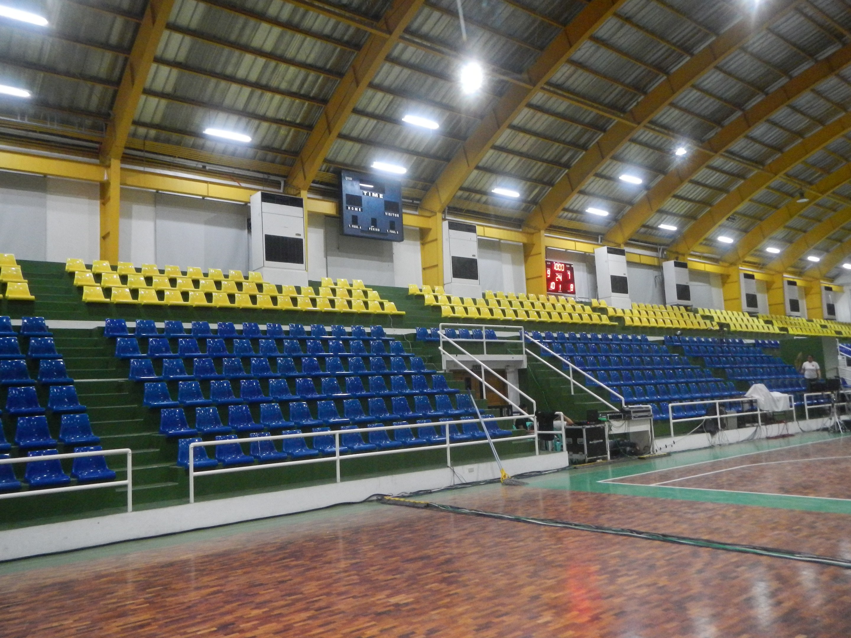 Große Sporthalle mit Holzboden, blauen und gelben Sitzplätzen, Geländern, Treppen mit Geländern, Wandbildschirmen, Deckenleuchten und einigen Menschen.