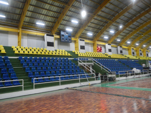 Große Sporthalle mit Holzboden, blauen und gelben Sitzplätzen, Geländern, Treppen mit Geländern, Wandbildschirmen, Deckenleuchten und einigen Menschen.