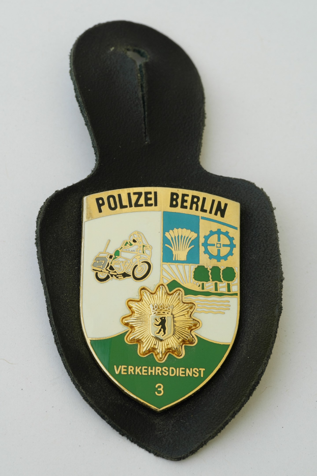 Ein schildförmiges Polizeibadget mit der Aufschrift "Polizei Berlin" auf einem blauen Hintergrund, einem weißen Rand, einem weißen Stern in der Mitte und schwarzer fetter Schrift auf einer Oberfläche.