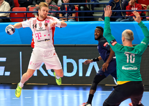 Eine Gruppe von Männern, die Handball auf einem Platz mit einem Ball in der Mitte spielen, während Zuschauer von Stühlen und Geländern in einem Stadion zuschauen, mit einem Schild im Hintergrund, auf dem "Futsal-Weltmeisterschaft 2019 - Bayern München vs. Paris Saint-Germain" steht.