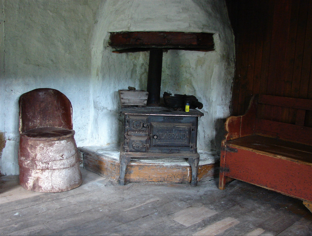 Altmodischer Holzofen in einem Raum mit einer hölzernen Bank auf der rechten Seite, einem Stuhl auf der linken Seite und einer Wand im Hintergrund.