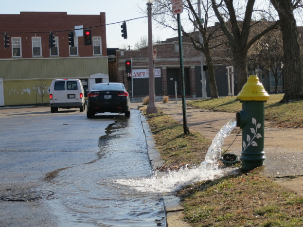 Ein Feuerhydrant auf dem Gehweg lässt einen Wasserstrahl frei, mit Fahrzeugen, Verkehrszeichen, Gebäuden, Grünflächen und Himmel im Hintergrund.