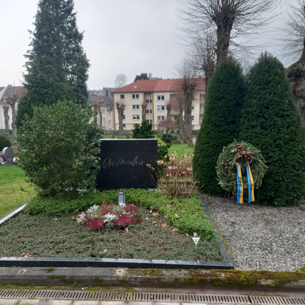 Ein Denkmal für die Opfer des Zweiten Weltkriegs steht in einem Park, geschmückt mit einem Kranz und umgeben von Grün, mit Gebäuden im Hintergrund.