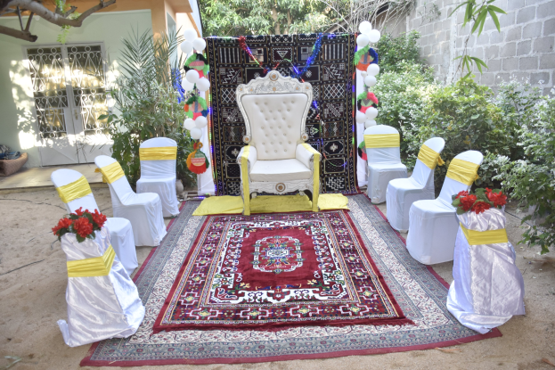 Hochzeitszeremonie-Aufbau im Garten mit weißen Stühlen, einem roten Teppich, Blumenbouquets, Dekorationsgegenständen, Ballons, Pflanzen, Bäumen, einem Gebäude mit Fenstern und einer Tür sowie einer Wand auf der rechten Seite.