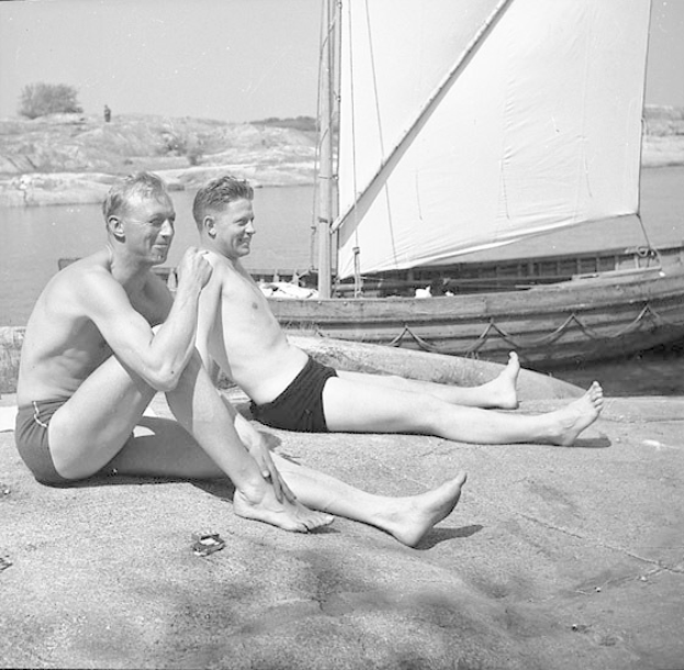 Zwei Männer in Badesachen sitzen am Strand neben einem Segelboot, mit blauem Wasser, grünen Bäumen und einem weißen Himmel im Hintergrund.
