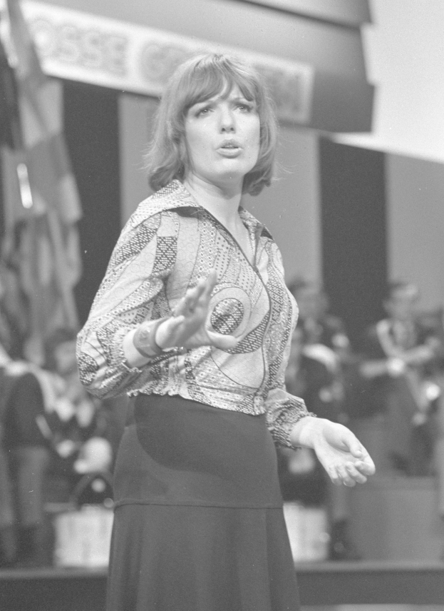 Schwarzes und weißes Foto einer Frau in einem weißen Hemd und schwarzer Hose auf einer Bühne mit einem ernsten Gesichtsausdruck, umgeben von einer Gruppe von Menschen und einer Wand im Hintergrund.