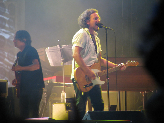 Ein Mann in einem weißen Hemd spielt Gitarre und singt in ein Mikrofon auf der B├╝hne, neben ihm spielt ein anderer Mann in einem schwarzen Hemd Gitarre; ein Piano, Kabel, Lautsprecher und Lichter sind im Hintergrund zu sehen.