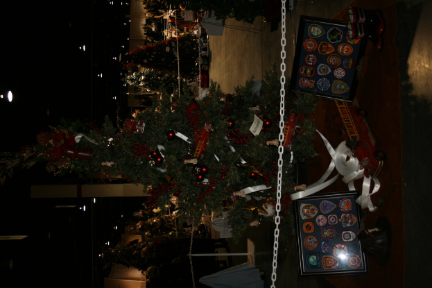 Ein Weihnachtsbaum mit Geschenken, Rahmen und Spielzeug auf einer Plattform, mit Menschen, zusätzlichen Weihnachtsbäumen, Ketten, Polen und Lichtern im Hintergrund.