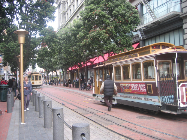 Eisenbahnschienen verlaufen durch eine Straße mit Zügen, die Text anzeigen, einer Person in der Nähe, kleinen Pfählen mit Seilen auf der linken Seite, Straßenlaternen, Bäumen, vielen Menschen, einem Müllcontainer auf der linken Seite und Gebäuden mit Leitern auf der rechten Seite.