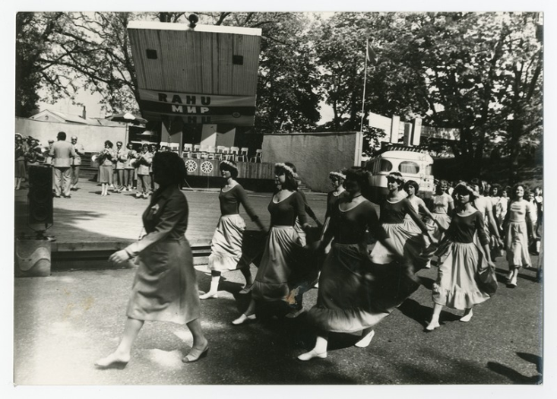 Eine Gruppe von Frauen, die auf der Straße tanzen, umgeben von einer Menge, einem Lautsprecher, einem Fahrzeug, einer Tafel mit Text, einer Fahnenstange, Gebäuden und einem bewölktem Himmel.
