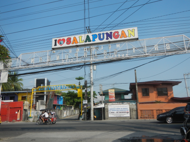 Eine Straßenszene mit Fahrzeugen, einem Bogen mit der Aufschrift "Iosalapungan", Strommasten, Gebäuden, Bäumen und einem Himmel im Hintergrund.