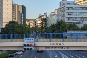 Eine Stadtstraße mit fahrenden Autos, eine Brücke mit Geländern und einem Schild, Verkehrsampeln, Laternen, Bäume und Gebäude mit Fenstern unter einem klaren Himmel.