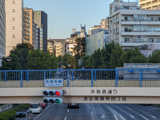 Eine Stadtstraße mit fahrenden Autos, eine Brücke mit Geländern und einem Schild, Verkehrsampeln, Laternen, Bäume und Gebäude mit Fenstern unter einem klaren Himmel.