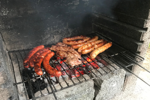 Grill mit Würstchen und Fleisch darauf, umgeben von brennender Kohle und Flammen, draußen auf Gras gesetzt.