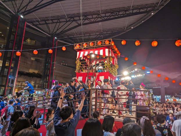 Eine Menschenmenge auf einer beleuchteten Bühne mit Laternen, einige halten Kameras, umgeben von einem Geländer, mit Bäumen und einem blauen Himmel im Hintergrund, möglicherweise während eines japanischen Festivals.