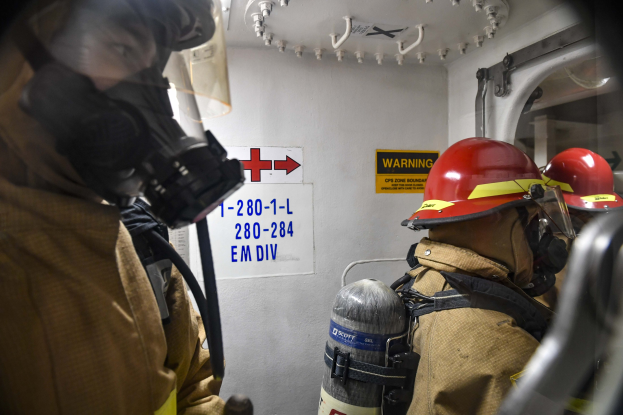 Zwei Feuerwehrleute in Schutzausrüstung stehen nebeneinander in einem Raum mit textgefüllten Tafeln an der Wand und Rohren darüber.