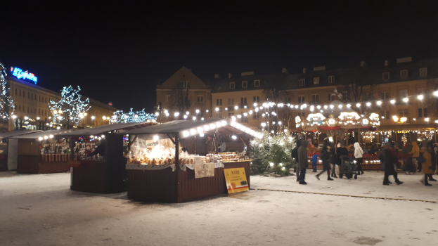 Ein nächtlicher Weihnachtsmarkt auf einer verschneiten Straße mit Menschen, Buden mit Lichtern, Pflanzen, Bäumen, Gebäuden, Schildern und einem bewölkten Himmel.
