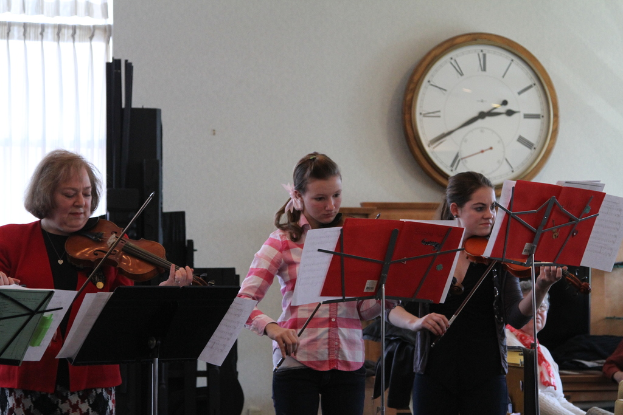 Drei Frauen auf der Bühne mit Geigen spielend, mit Notenpulten und Noten vor sich, während ein Zuschauer im Vordergrund sitzt; eine Uhr ist an der weißen Wand im Hintergrund angebracht, und ein weißer Vorhang ist links im Bild zu sehen.