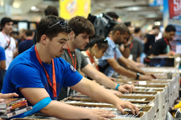 Eine Gruppe von Menschen mit Ausweisen packt Bücher in Kisten, mit einem Banner oben rechts und anderen Menschen im Hintergrund.