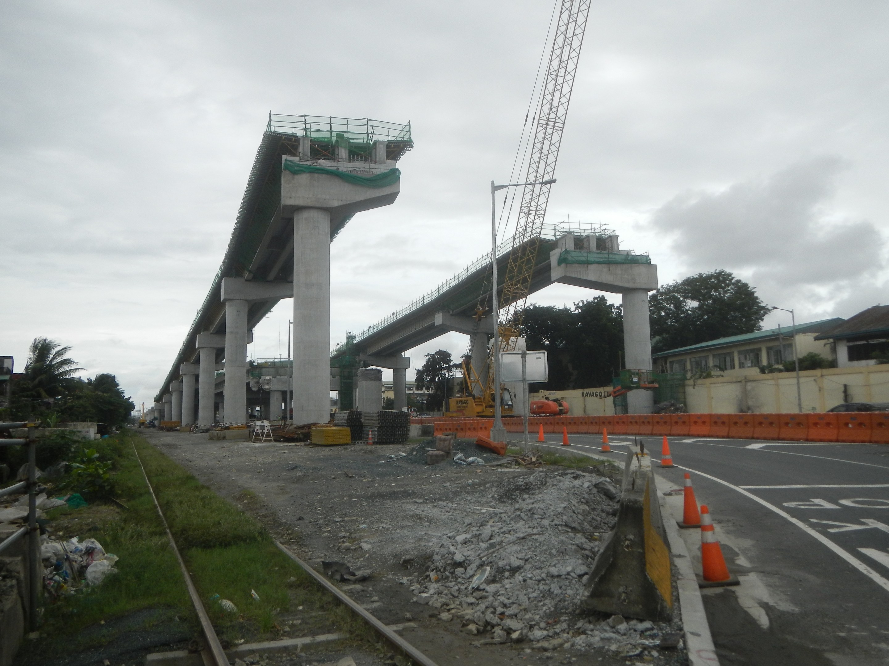 Baustelle mit einer Brücke im Hintergrund, Straße mit Absperrkegeln markiert, Eisenbahnschienen auf der linken Seite, verstreute Steine und Gras, Bäume und Gebäude säumen die Straße und ein bewölkter Himmel.