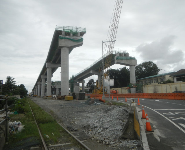 Baustelle mit einer Brücke im Hintergrund, Straße mit Absperrkegeln markiert, Eisenbahnschienen auf der linken Seite, verstreute Steine und Gras, Bäume und Gebäude säumen die Straße und ein bewölkter Himmel.
