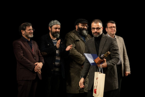 Eine Gruppe Männer steht auf einer Bühne, einer hält eine Tasche und eine Trophäe, mit dunklem Hintergrund, der die Eröffnungsnacht des Berliner Filmfestivals vermuten lässt.