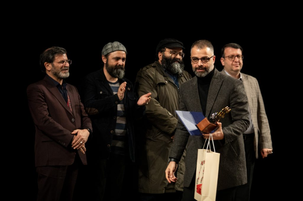 Eine Gruppe Männer steht auf einer Bühne, einer hält eine Tasche und eine Trophäe, mit dunklem Hintergrund, der die Eröffnungsnacht des Berliner Filmfestivals vermuten lässt.