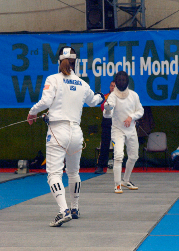 Zwei Fechter in weißen Kostümen messen sich bei den Olympischen Spielen, jeder hält ein Schwert in der Hand, mit einem blauen Banner und Zuschauern im Hintergrund.