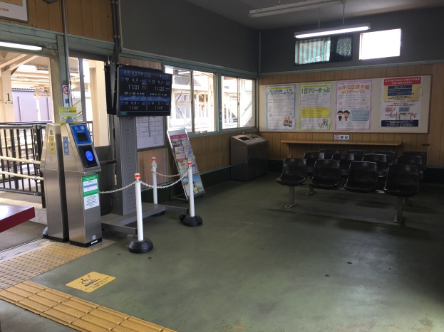 Ein belebter Bahnhofseingang mit Reihen von Stühlen, einem Service-Schalter, Ticketautomaten, Kettengliedern, Informationsanzeigen, Wandplakaten, Deckenleuchten und Geländern.